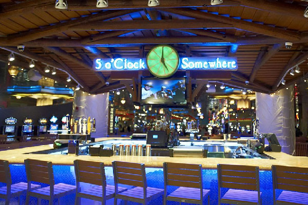 A brightly lit bar with a wooden canopy and a neon “5 o’Clock Somewhere” sign above the counter.