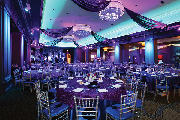 Elegant banquet hall set for a wedding with round tables, chandeliers, and purple lighting.
