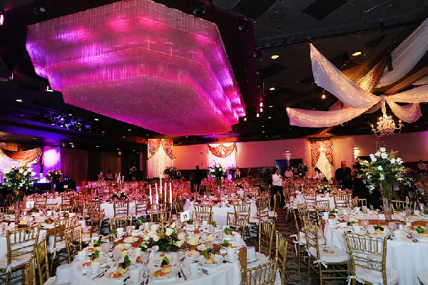 An elegant banquet hall set with round tables, gold chairs, floral centerpieces, and dramatic pink and gold ceiling décor.