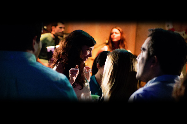 A group of people socializing in a venue, with one woman in focus holding a drink and dancing among others.