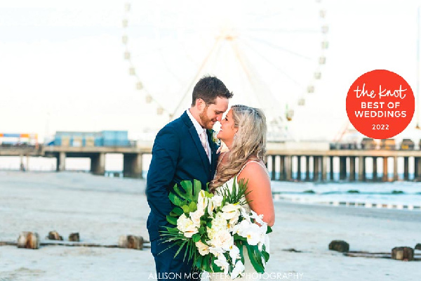 Couple in wedding attire holding a bouquet and smiling at each other on a beach with the boardwalk in the background.