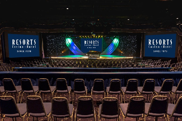 Theater-style auditorium with rows of seats facing a lit stage and large screens.