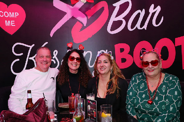 Four people sit together at a bar table, smiling at the camera with drinks in front of them and heart-themed accessories.