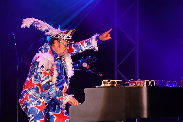 Elton John impersonator in a star-spangled suit and feathered hat pointing while playing a piano on stage.